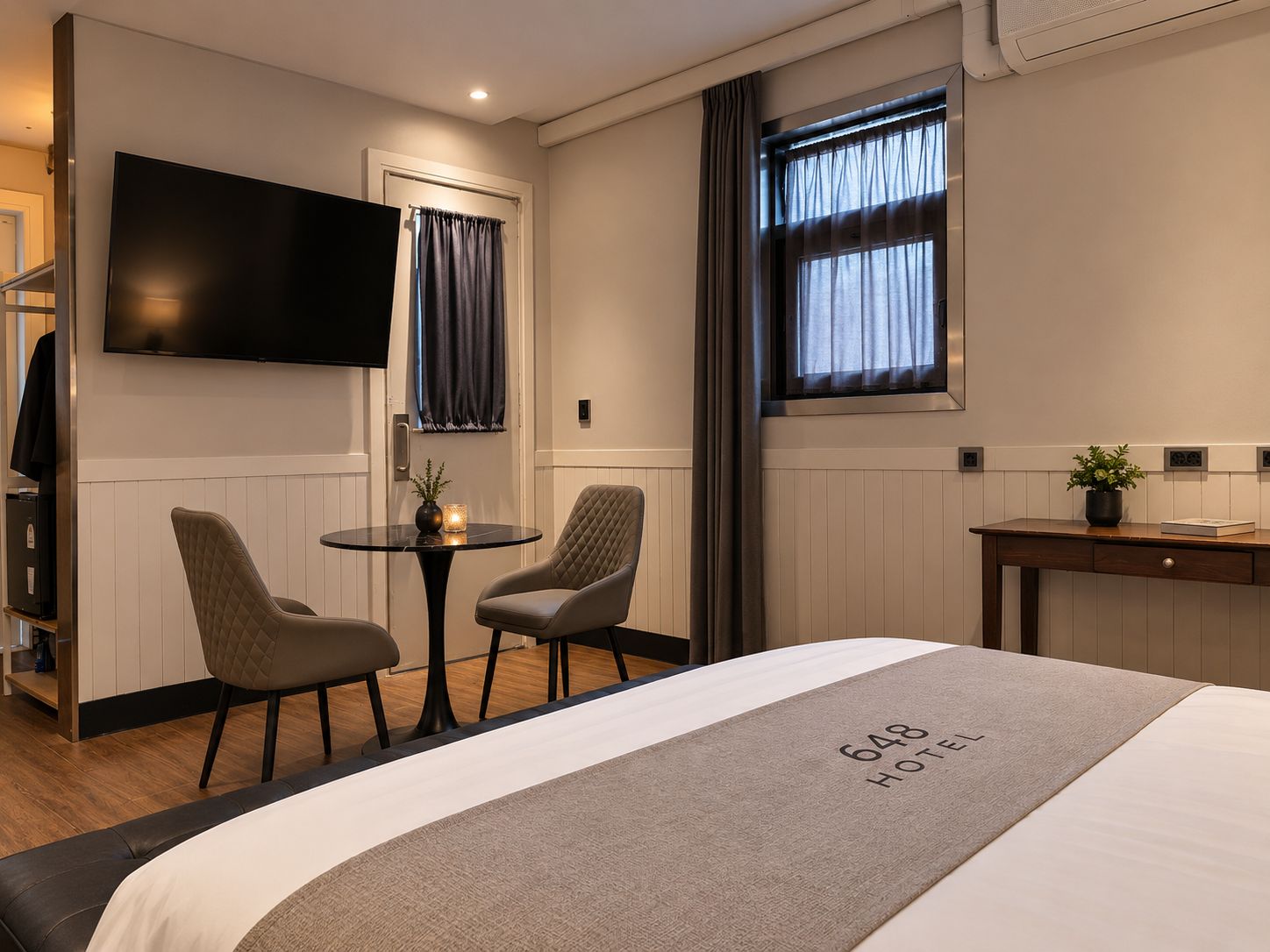 A hotel room with a bed, a TV mounted on the wall, a small table with chairs, and a window with curtains.