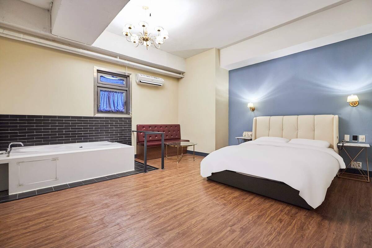 A modern hotel room with a large bed, a separate bathroom with a bathtub, a red leather sofa, and a TV mounted on the wall.