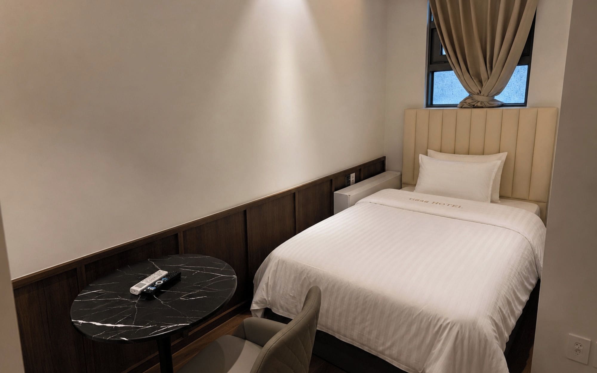 A hotel room with a bed, a bedside table, a remote control, and a window with curtains.