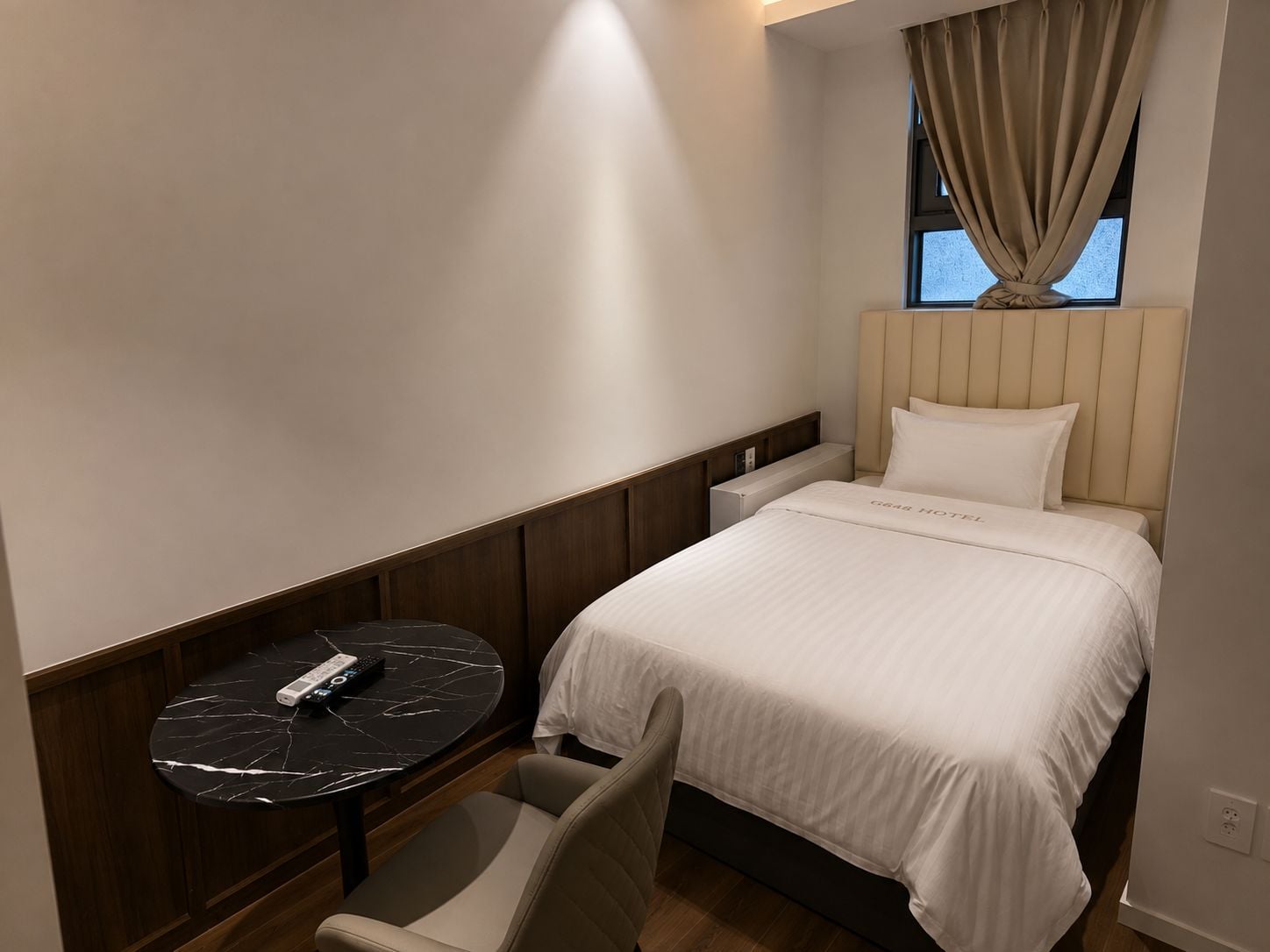 A hotel room with a bed, a small round table with a remote control, a chair, and a window with curtains.
