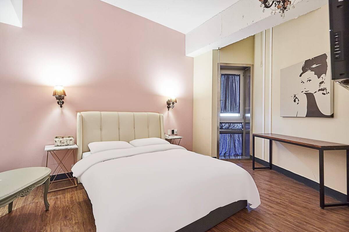 A bedroom with a large bed, two bedside tables, a nightstand, a chandelier, and a painting on the wall.