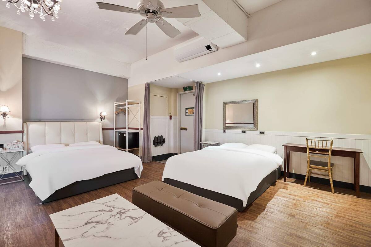 A hotel room with two beds, a desk, a chair, a ceiling fan, and a mirror.