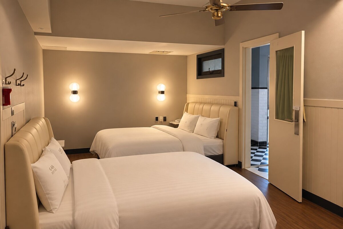 A hotel room with two beds, a ceiling fan, wall-mounted lights, and a door leading to a hallway.