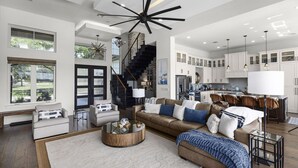 A spacious living room with a large sofa, a coffee table, and a fireplace. There is a kitchen area with white cabinets and a dining table. - Whispering Waters - Lakefront Luxe, Pool + Hot Tub + Fire Pit + Outdoor Kitchen + 2 Master Suites (Canyon Lake)