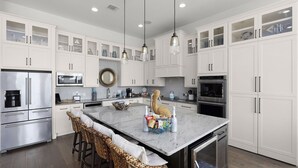 A modern kitchen with white cabinetry, stainless steel appliances, a marble island, and wicker bar stools. - Whispering Waters - Lakefront Luxe, Pool + Hot Tub + Fire Pit + Outdoor Kitchen + 2 Master Suites (Canyon Lake)
