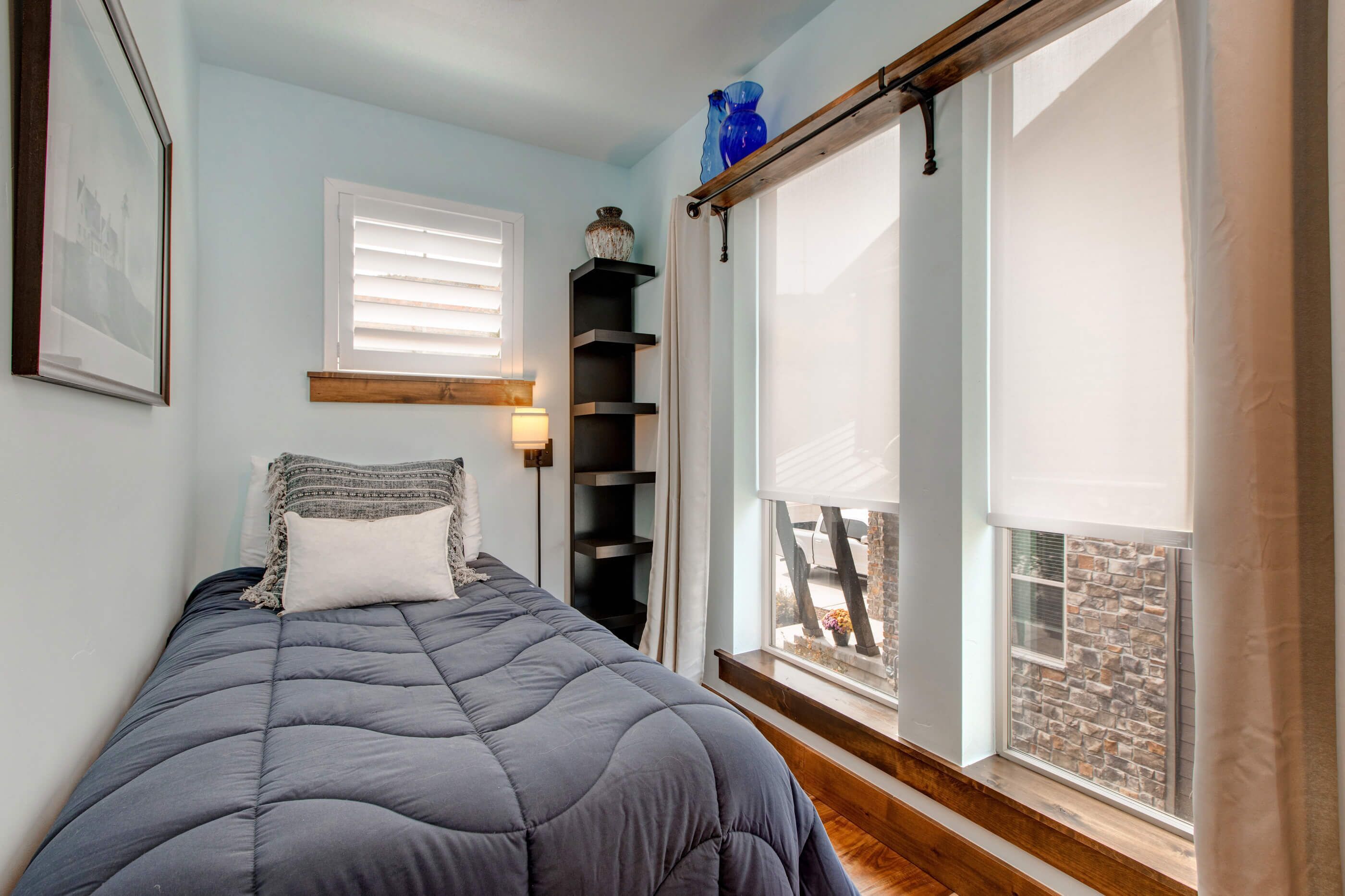 Une chambre à coucher avec un lit, une fenêtre avec des stores, une étagère ornée d’objets décoratifs et une vue sur l’extérieur.