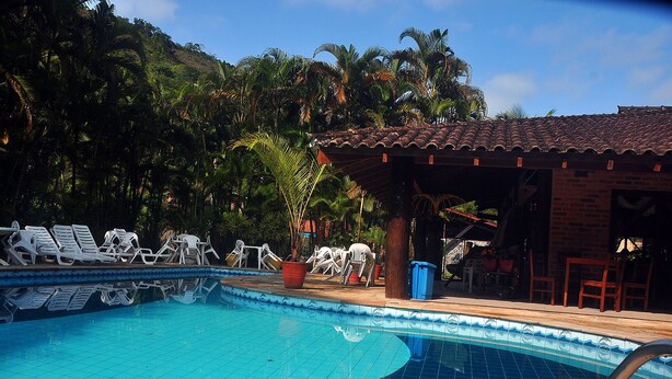 Uma piscina com espreguiçadeiras, área coberta e vegetação exuberante ao fundo.
