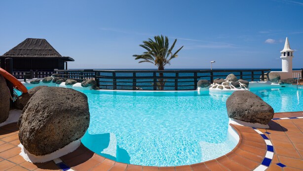 Una piscina azul cristalina con borde de azulejos, rodeada de rocas y una terraza de madera.