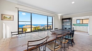 A spacious dining area with a large table and chairs, a bookshelf, and a view of the ocean. - Montague Views Kianga (KIANGA)