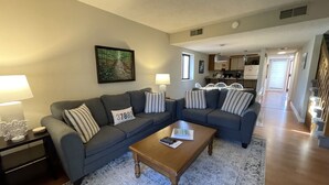 Ein Wohnzimmer mit zwei grauen Sofas, einem Holztisch und einem Teppich. An der Wand hängt ein Gemälde und im Hintergrund ist eine Küche zu sehen.