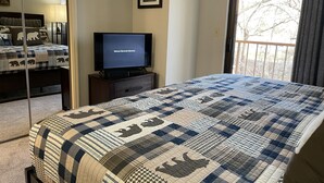 Ein Schlafzimmer mit Bett, Fernseher auf einem Schrank, einem gerahmten Bild an der Wand und einem Fenster mit Blick auf Bäume.