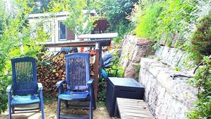 An outdoor seating area with two chairs and a table, surrounded by dense foliage and a stone wall.