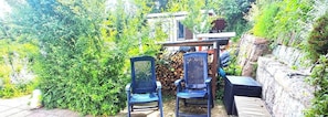 Un espace extérieur aménagé avec deux chaises et une table, entouré d’une végétation luxuriante et d’un mur de pierre.
