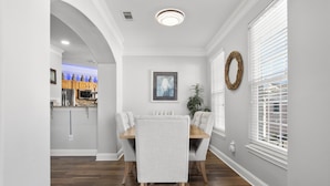 Un coin repas avec une table et des chaises en bois, un tableau au mur et une fenêtre avec des stores.
