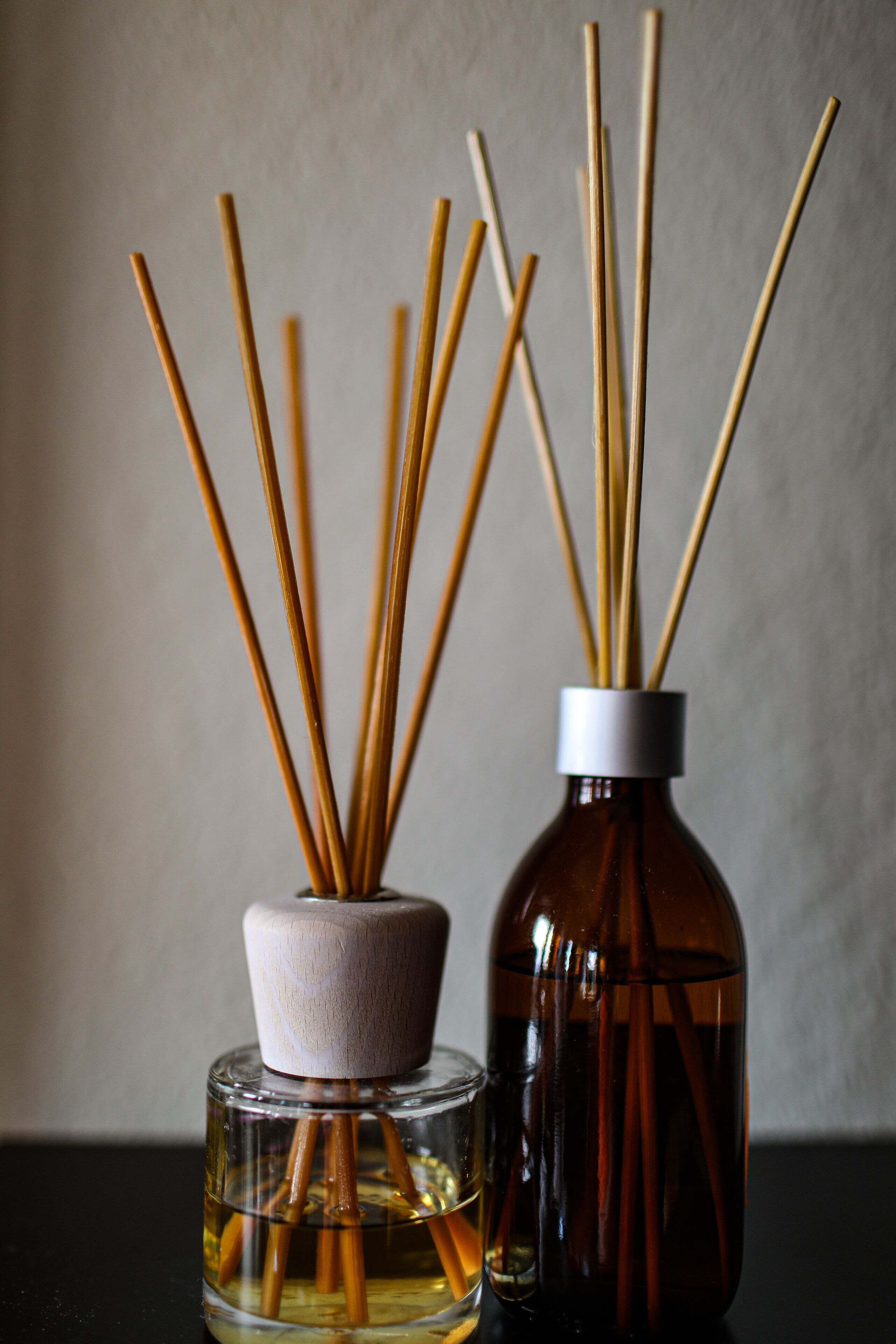 Two bottles with reed diffusers, one in a clear glass and one in a brown bottle, placed on a surface.