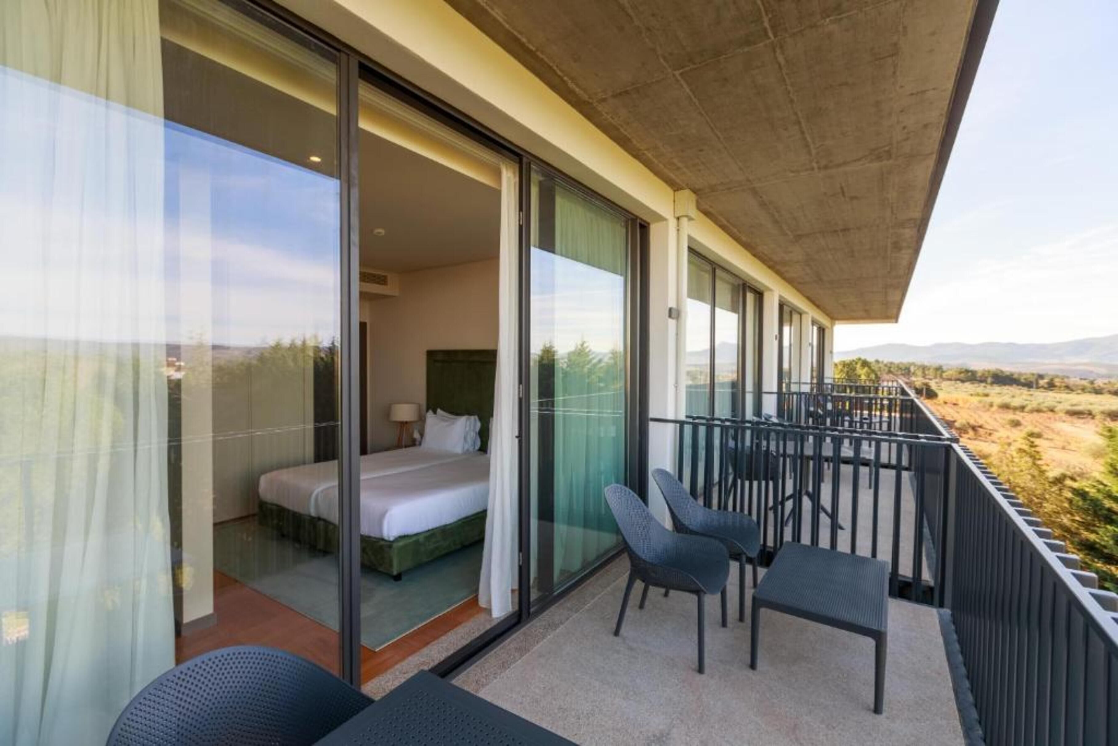 A balcony with a bed, two chairs, and a view of the countryside.
