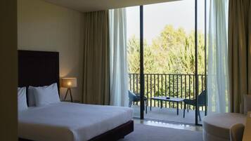 A hotel room with a large bed, a bedside table with a lamp, a balcony with a table and chairs, and a view of trees.