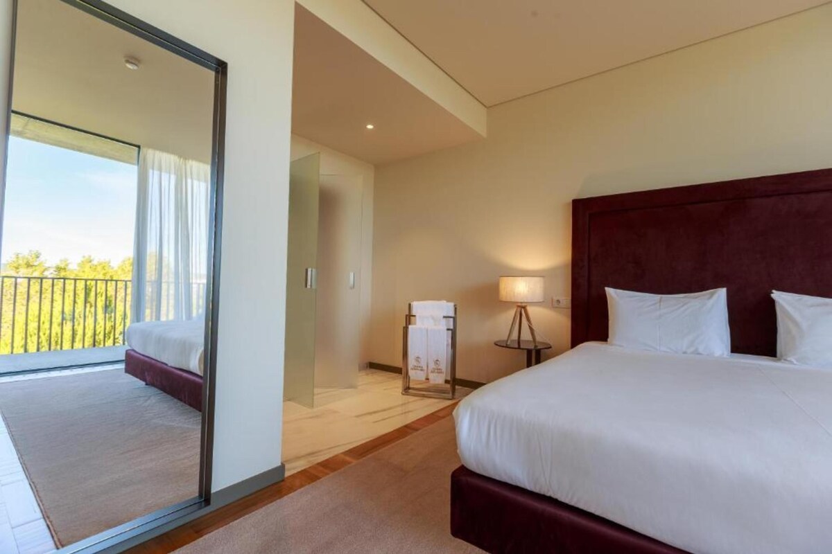 A hotel room with a large bed, a bedside table with a lamp, a view of the balcony, and a door leading to another room.