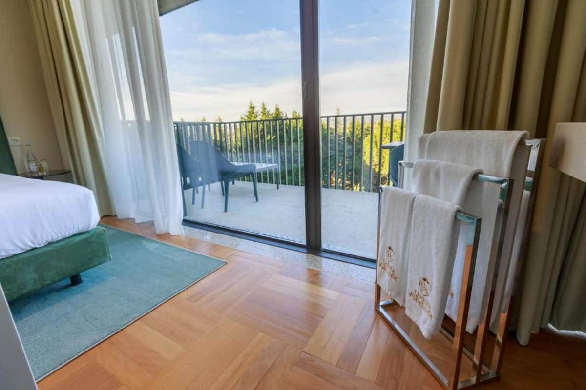 A hotel room with a bed, a green sofa, a balcony with chairs, and a towel rack.