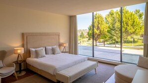 A modern bedroom with a large bed, bedside tables, a bench, and a view of a green landscape through large windows. - Olive Nature - Hotel & SPA (Valpacos)