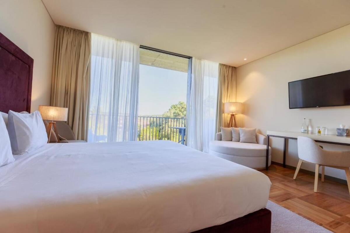 A hotel room with a large bed, two bedside tables, a desk, a chair, a TV, and a balcony with curtains.