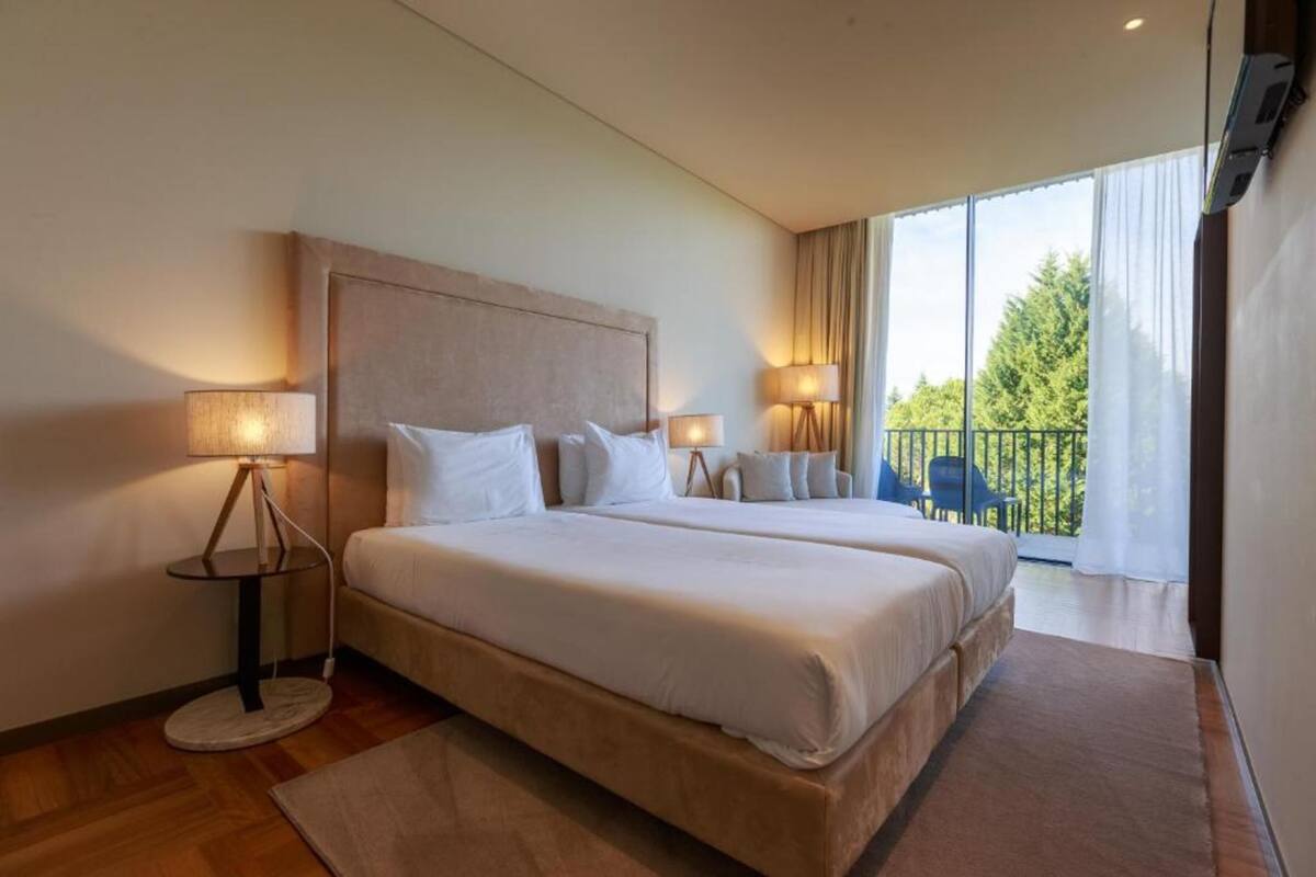 A hotel room with a large bed, two bedside lamps, a view of the outdoors through a balcony.
