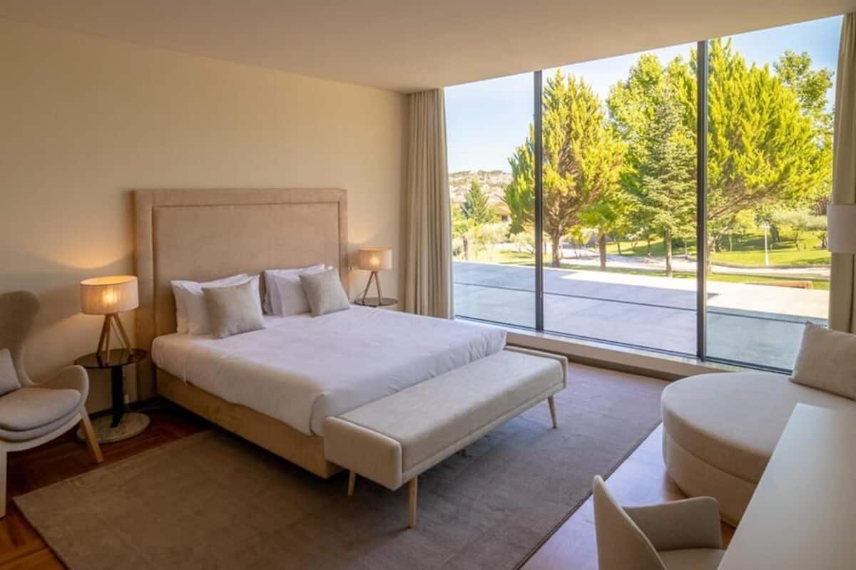 Habitación de hotel moderna con una cama grande, una zona de estar con sofá, una silla y vistas a un paisaje verde a través de amplios ventanales.
