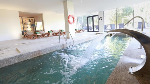 An indoor swimming pool with a waterfall feature, surrounded by lounge chairs and large windows. - Olive Nature - Hotel & SPA (Valpacos)