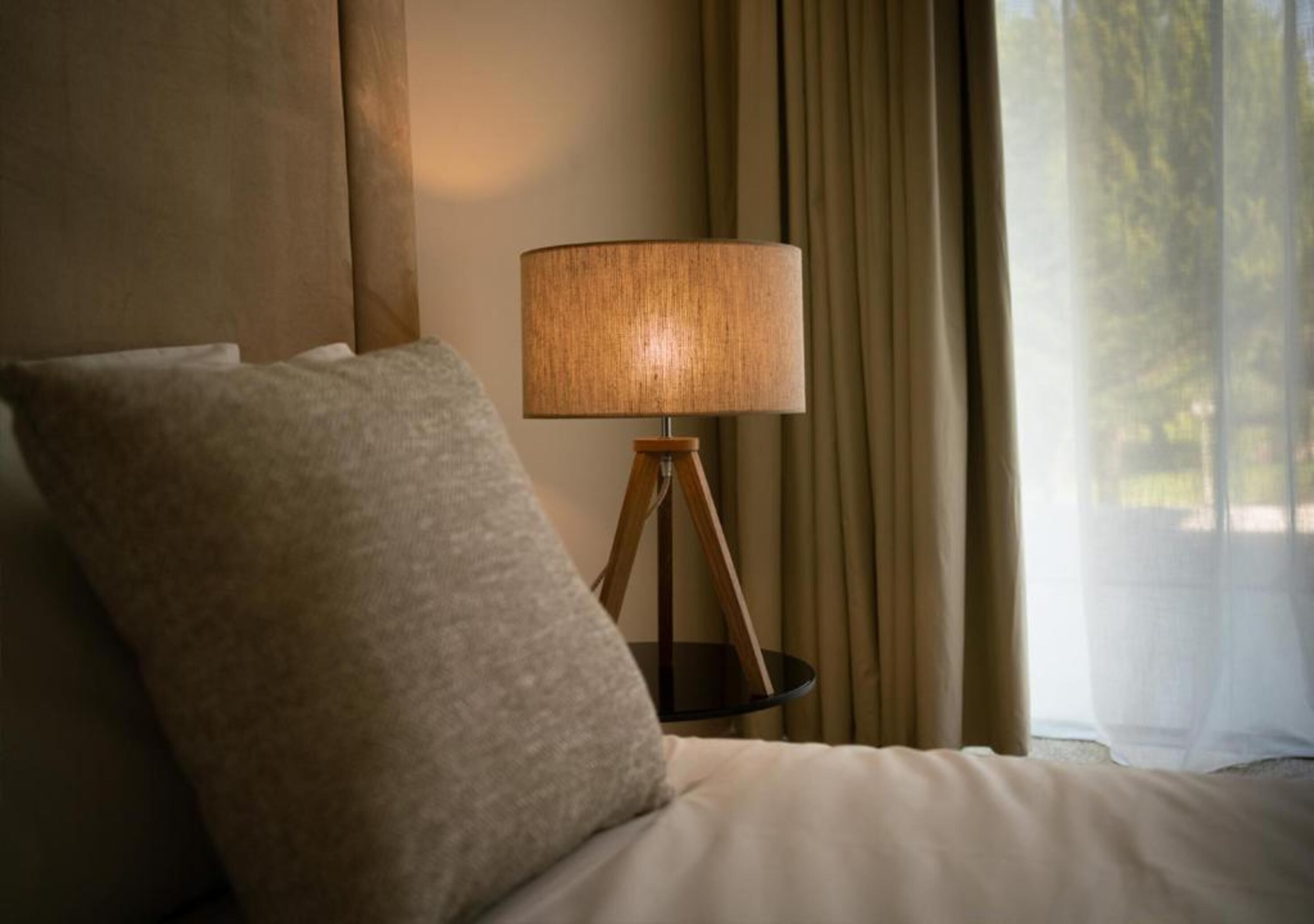 A bed with a single pillow, a bedside lamp on a wooden stand, and a window with curtains.