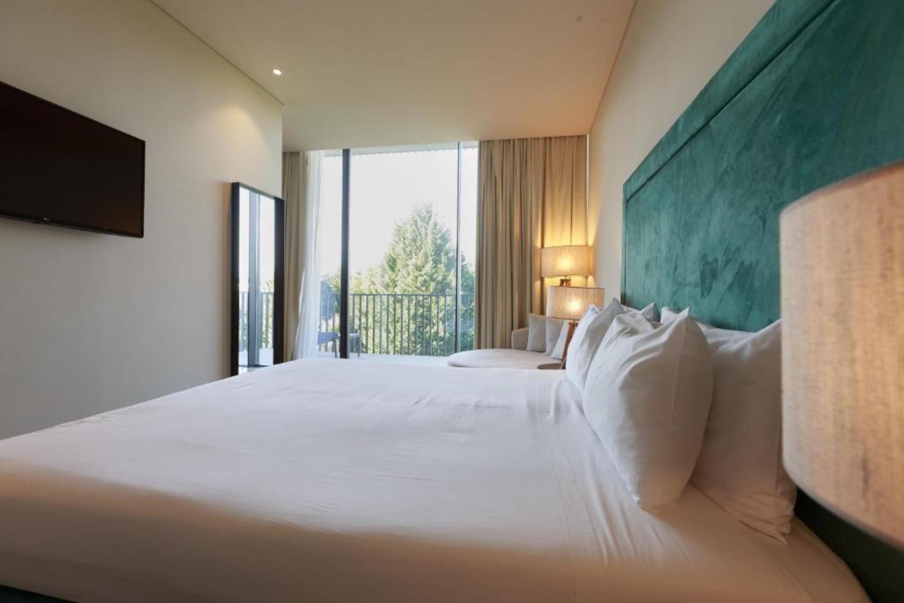 A hotel room with a large bed, a view of the outdoors, a television mounted on the wall, and a lamp on the bedside table.