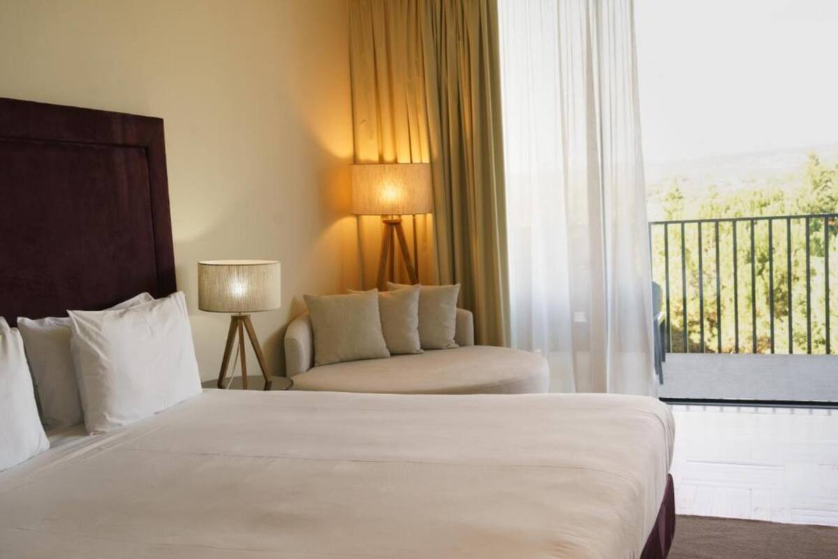 A hotel room with a large bed, a bedside table with a lamp, and a view of greenery through the window.