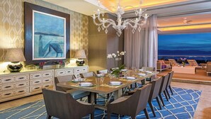 Une salle à manger avec une longue table, des chaises et un lustre. Un tableau orne le mur, et de grandes fenêtres offrent une vue sur l’océan.