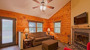 Interior acogedor de una cabaña de madera con chimenea, un sofá, un sillón rojo y un ventilador de techo.