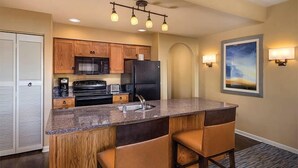 A kitchen with wooden cabinets, a black refrigerator, a microwave, a stove, and a granite countertop. There are two bar stools and a painting on the wall. - Phoenix | Desert Golf Retreat | Pool, Dining & Sunsets (Phoenix)