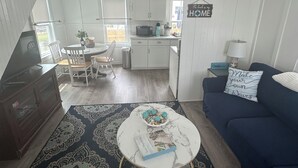 A living room with a blue sofa, a coffee table, and a patterned rug. There is a kitchen in the background with white cabinets and a toaster. - Quaint Cottage Vacation (Old Orchard Beach)