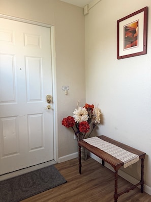 A hallway with a white door, a bench with a woven seat, a vase with flowers, and a framed picture on the wall.