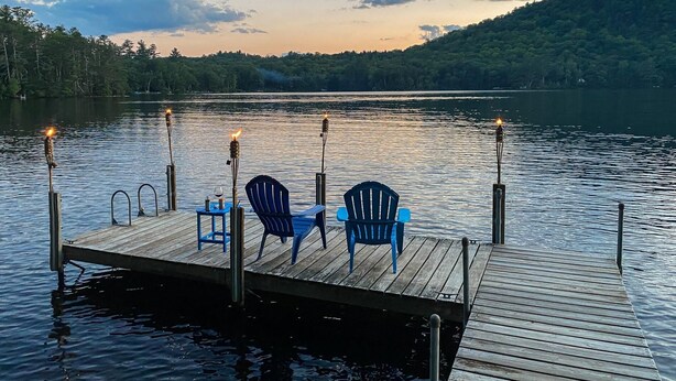 Une jetée en bois, avec deux chaises bleues et une table, entourée d’une étendue d’eau calme et d’une colline boisée.
