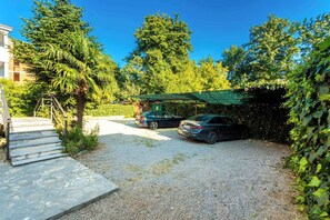 Uma estrada de cascalho leva a um carro estacionado sob uma copa verde, com uma escadaria de pedra à esquerda e um edifício ao fundo.