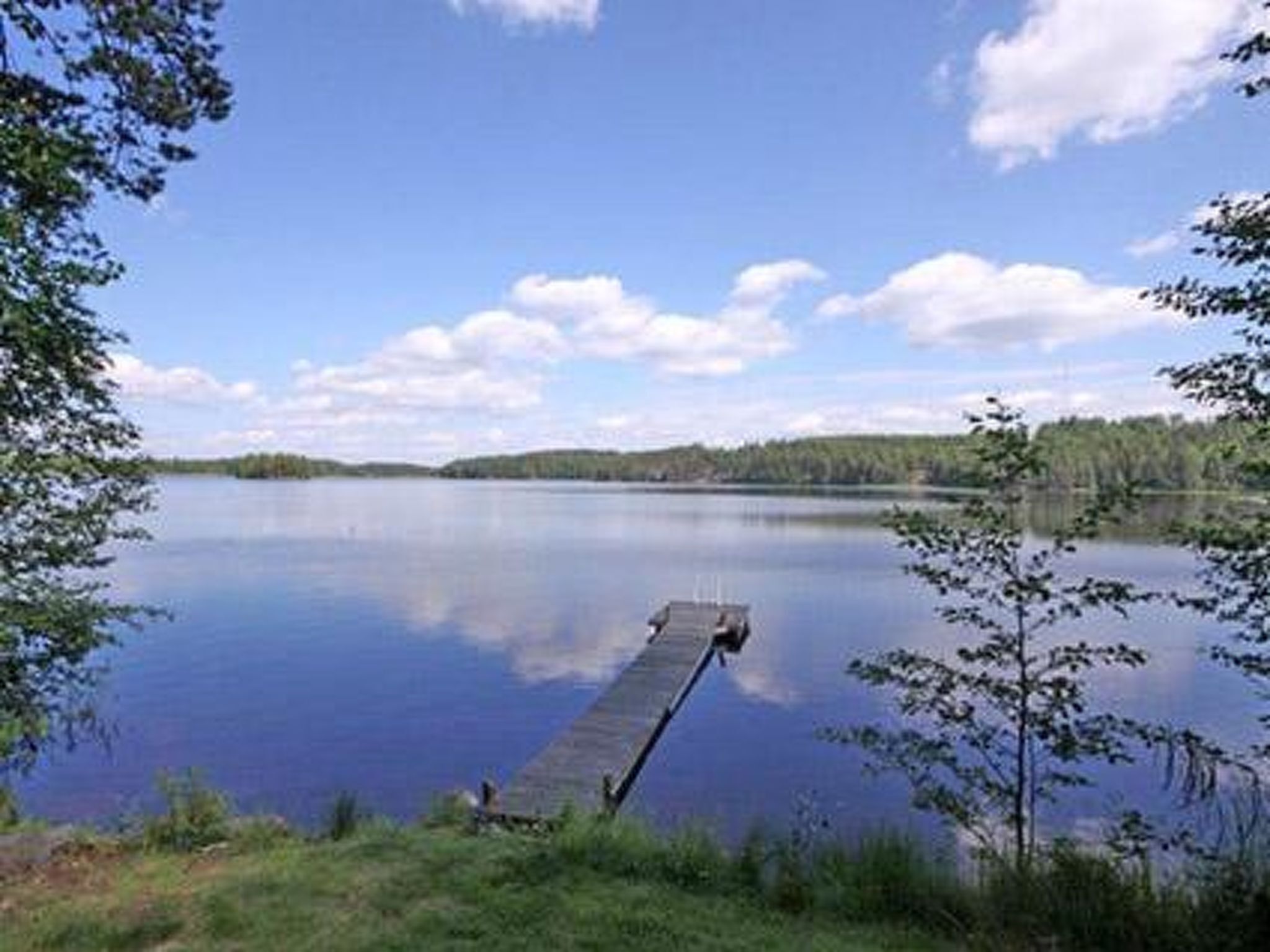 A calm lake with a wooden dock extending into the water, surrounded by trees and clear skies.