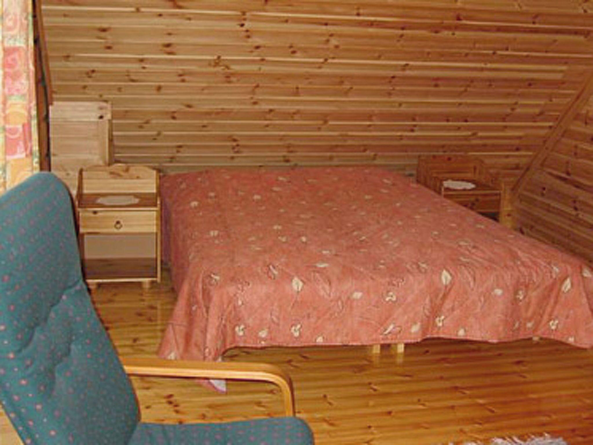 Una habitación pequeña y acogedora, con techo de listones de madera, una cama individual con colcha estampada, una mesita de noche de madera y una silla.