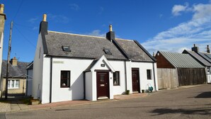 A white building with a dark roof, a chimney, and a small porch with a door and windows. - 4-bed Cottage in Portknockie, Near Cullen, Moray (Buckie)