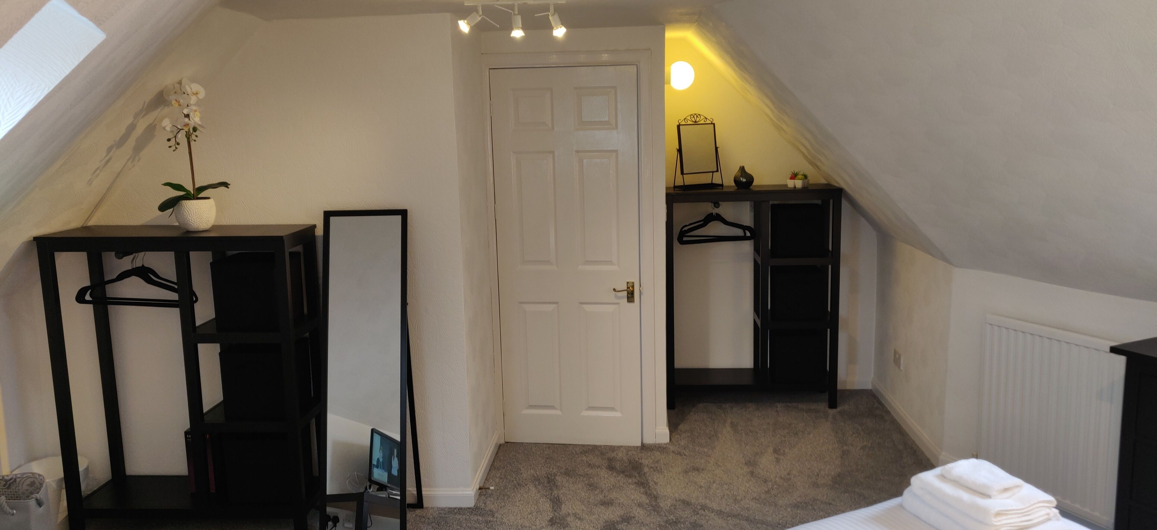 A small, well-lit attic bedroom with a white door, a black shelving unit with a hanging rack, a small table with a plant, and a radiator.