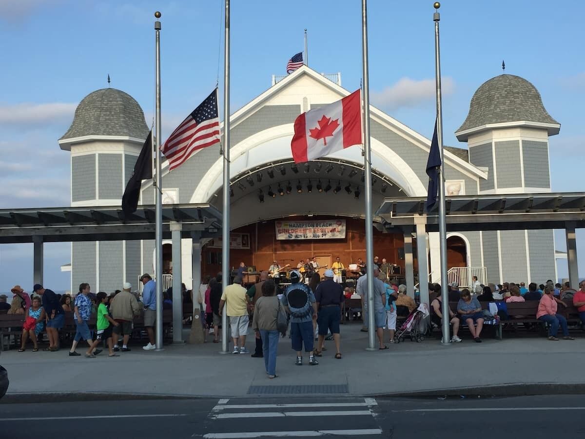 A covered outdoor area with a stage and audience seating, flags, and a clear sky.