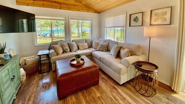 A cozy living room with a wooden ceiling, a beige sectional sofa, a red ottoman, a small side table, and a floor lamp.