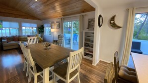 A spacious living area with a wooden dining table, white chairs, a sofa, and a view of the outdoors through large windows.