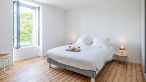A bedroom with a bed, a small table with a lamp, and a window with a view of greenery.