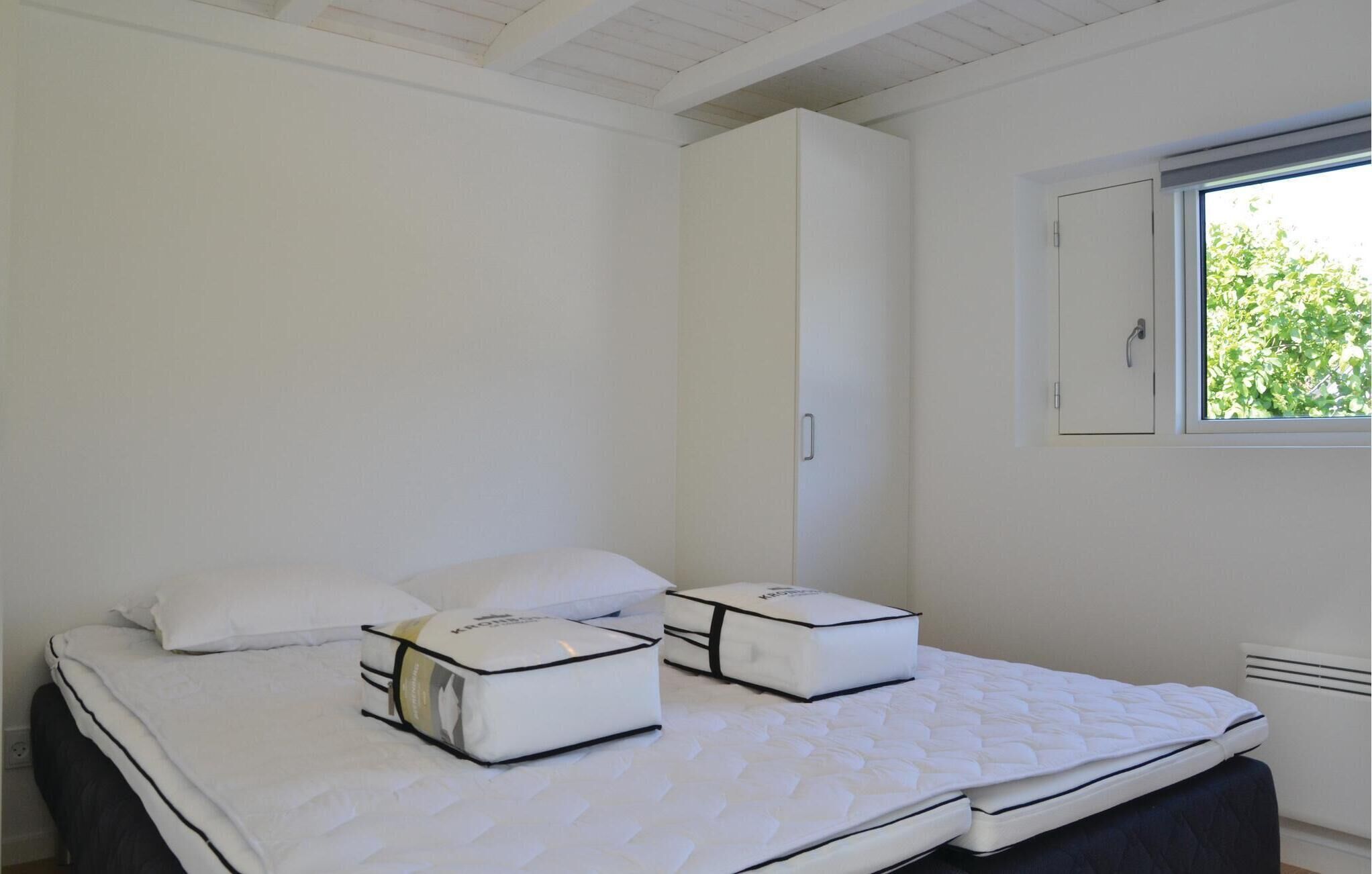 A bedroom with two beds, each with a white mattress and matching pillows, a window with a view of greenery, and a white wardrobe.