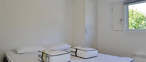 A bedroom with two beds, each with a white mattress and matching pillows, a window with a view of greenery, and a white wardrobe.