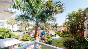A balcony with a view of a swimming pool, palm trees, and a thatched-roof structure. - Charming Studio Beach Getaway. Beachfront Pool, Near Restaurants, Activities (St. Pete Beach)