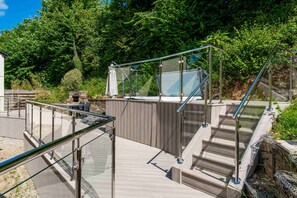 Un espace extérieur clos par des parois de verre, doté d’une terrasse en bois, d’un mur en pierre et d’un escalier menant à un niveau supérieur.